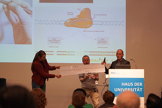 Three people on the stage in front of a presentation cutting a paper with a DNA sequence printed on it